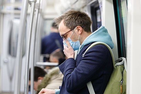 A masked man on a train is holding his mouth, looking down from his seat. 